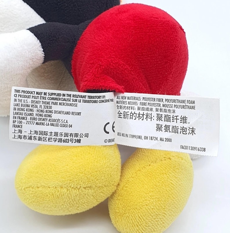K&ouml;nnte beinhalten: Ein Pl&uuml;schtier mit roten Shorts, gelben Schuhen sowie schwarzen und wei&szlig;en Stoffteilen. Ein wei&szlig;es Stoffetikett ist angebracht, das gedruckten Text auf Englisch, Franz&ouml;sisch und Chinesisch zu den Materialien und Vertriebsorten enth&auml;lt.
