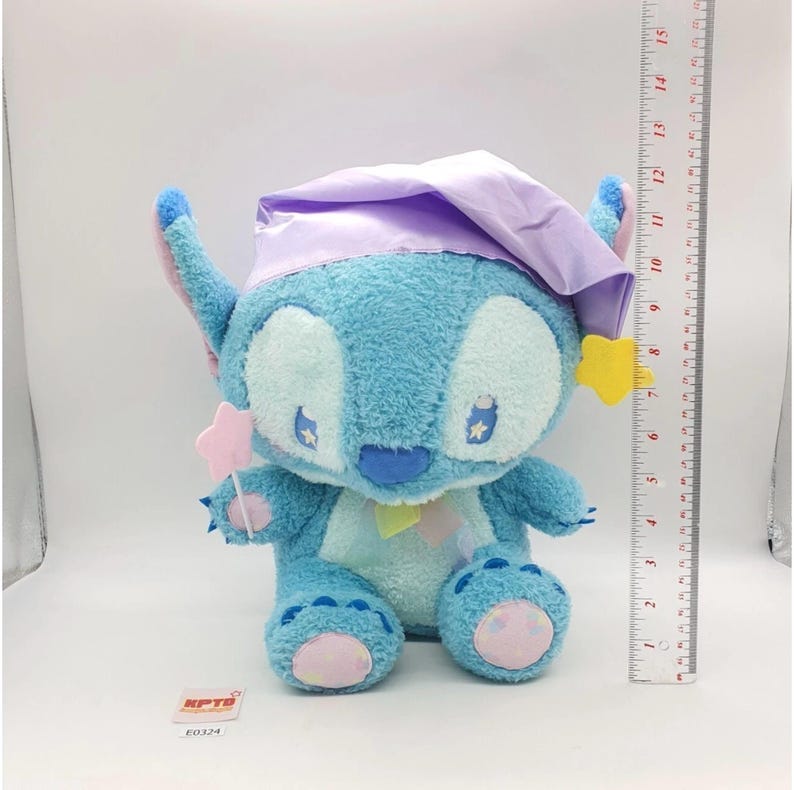 May include: A plush blue Stitch toy wearing a lavender nightcap and holding a pink star wand. The toy has yellow star accents and is approximately 10 inches tall, with a ruler in the background.