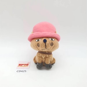 以下が含まれることがあります： ピンクの帽子と茶色の襟をつけたぬいぐるみキャラクター。おもちゃは、タン色の体、濃い灰色の足、青い鼻をしています。キャラクターは、頬に渦巻き模様があり、真剣な表情をしています。