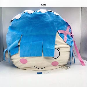 May include: A plush, round pillow shaped like a cartoon character's face. The pillow features blue hair with white accents, pink accents, and a cream-colored face with black and pink facial details. The pillow is on a white surface.