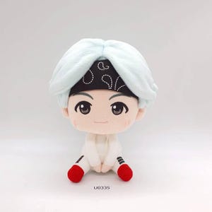 May include: A plush doll with light blue hair and a black bandana with white designs. The doll has large, expressive eyes, a small smile, and is wearing a white outfit with red accents on the feet. The tag reads "U0335".