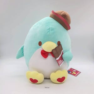 Könnte beinhalten: Ein Plüschpinguin in Türkis, Weiß und Gelb, mit braunem Hut und roter Fliege. Der Pinguin hält einen Schokoriegel mit einem "Love Chocolate"-Etikett. Die Füße des Pinguins haben rote Herzakzente.