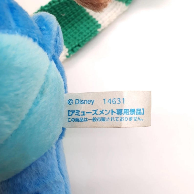 Puede incluir: Un peluche azul con una etiqueta de tela blanca que muestra el texto Disney 14631 y caracteres japoneses. El juguete tiene una textura suave y est&aacute; unido a una correa tejida de rayas verdes y blancas que sobresale de la parte superior.