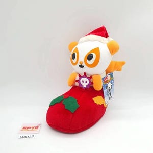 May include: A plush toy featuring a white and orange character wearing a red Santa hat, sitting inside a red Christmas stocking decorated with green holly leaves. The character has a purple gear-shaped emblem with a white skull on its chest.