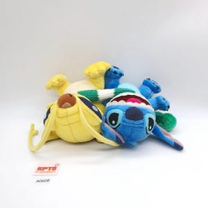 Puede incluir: Dos peluches que representan un personaje amarillo con antenas largas y un personaje azul con orejas grandes y boca ancha. Una bufanda de punto a rayas verdes y blancas envuelve ambas figuras. Una peque&ntilde;a etiqueta blanca muestra el texto KPTD A0608.
