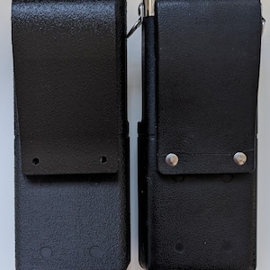 May include: Two black leather holsters with silver accents and metal rivets. The holsters have a textured surface and are designed to hold a radio or other device.