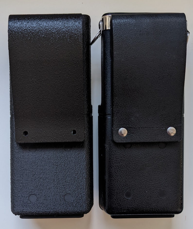 May include: Two black leather holsters with silver accents and metal rivets. The holsters have a textured surface and are designed to hold a radio or other device.