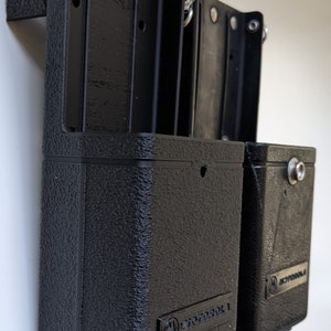 May include: Two black plastic radio holsters with the Motorola logo on the front. The holsters are mounted on a white wall.