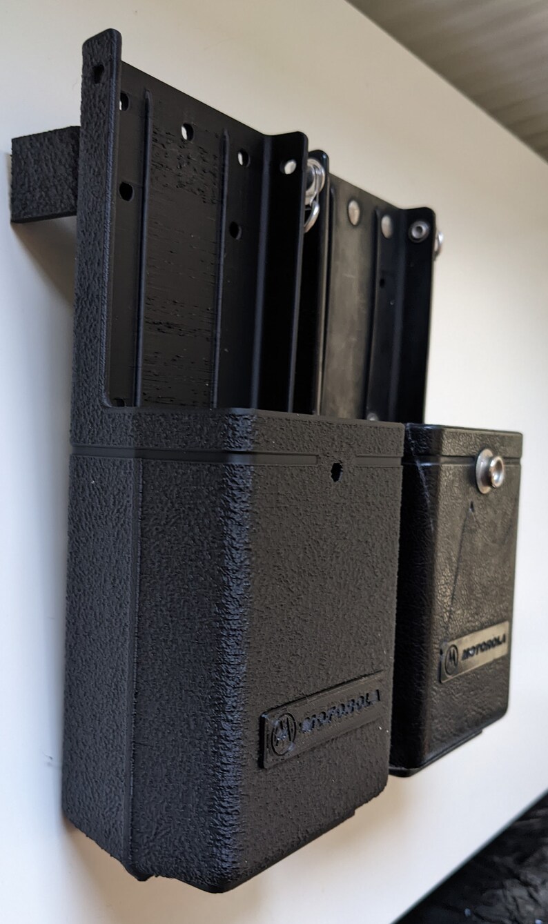 May include: Two black plastic radio holsters with the Motorola logo on the front. The holsters are mounted on a white wall.
