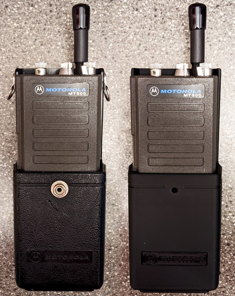 May include: Two black Motorola MT 600 two-way radios in black holsters. The radios have a black antenna and silver buttons.