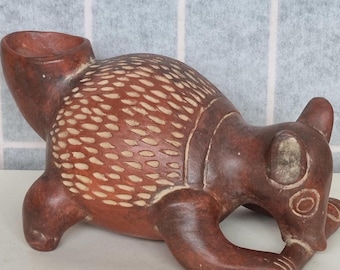 May include: A brown ceramic figurine of an animal with white markings. The animal is in a crouching position with its head down, as if it is sniffing the ground. The figurine has a textured surface and a small hole in the top of its head.