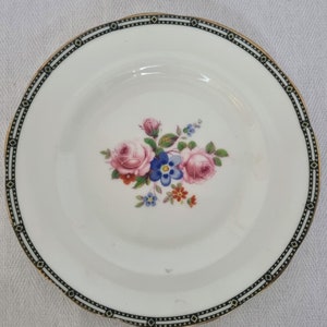 Puede incluir: Un plato de cerámica blanco con un borde negro y dorado. El plato presenta un diseño floral con flores rosas, azules y amarillas.