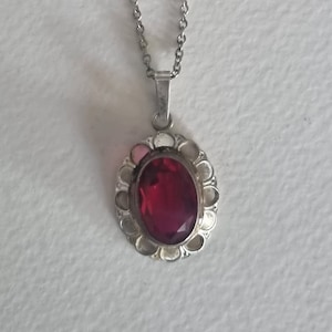 May include: A silver-toned necklace with a pendant featuring a large, oval, ruby-red gemstone. The pendant is surrounded by a decorative, scalloped frame. The chain is delicate and hangs against a white background.