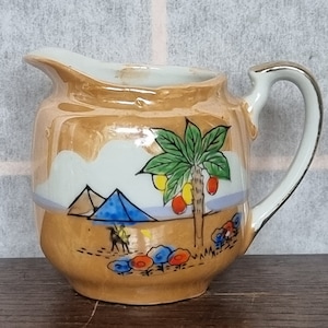 Può includere: Un piccolo creamer in ceramica marrone con un manico dorato. Il creamer è decorato con una scena dipinta di un paesaggio desertico con palme, piramidi e un cammello.