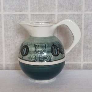 May include: A white ceramic pitcher with a green and black design. The pitcher has a round body and a handle. It features a crest and the word "JERSEY" on the front.