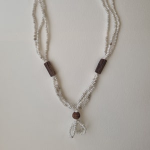 May include: A white beaded necklace with wooden accents. The necklace has three strands of beads and a wooden tassel at the bottom.