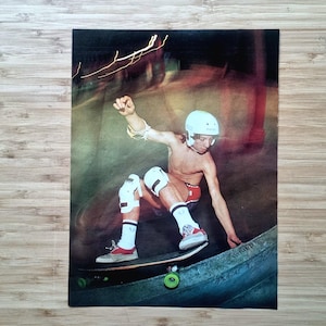 Puede incluir: Fotografía vintage de un skater en acción en una rampa. El skater lleva un casco blanco, rodilleras y pantalones cortos rojos. La imagen tiene una estética retro con movimiento borroso y un fondo oscuro.