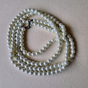May include: A white pearl necklace with a simple clasp. The necklace is made of multiple strands of pearls and is coiled on a light beige surface.