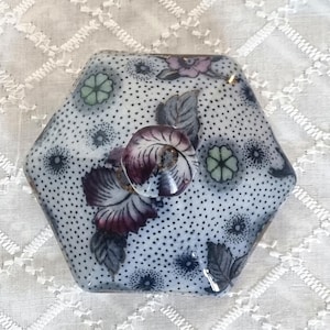 May include: Hexagonal ceramic lid with a floral design. The lid features a white background with small black dots, purple and pink flowers, and a central knob. The knob and flowers have a glossy finish.