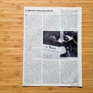 May include: A newspaper page from "Le Monde" with the headline "Le Monde's Changing World." The page features articles and a black and white photo of people working with the newspaper. The text is in black ink on a white background.