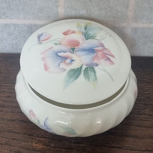 Può includere: Una scatola portaoggetti in ceramica bianca con un coperchio che presenta un motivo floreale di fiori rosa, blu e gialli. La scatola ha un bordo a festoni e un motivo floreale sul fondo.