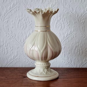 May include: A cream-coloured ceramic vase with a ruffled top and a bulbous body. The vase has a gold band around the neck and a decorative base. The body features vertical petal-like designs with a hint of pink. The vase is displayed on a wooden surface.