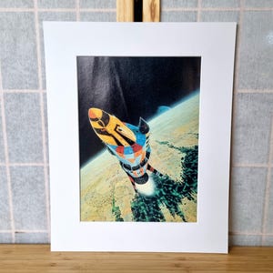 May include: A colorful rocket ship illustration against a backdrop of space and Earth. The rocket has a yellow, blue, and red design. The artwork is framed in a white mat.