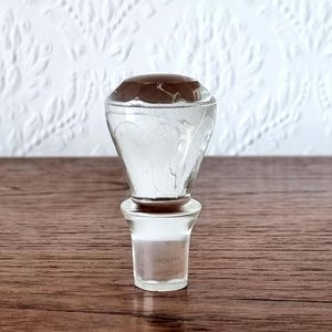Puede incluir: Un tapón de botella de vidrio transparente con una parte superior marrón facetada. El tapón tiene forma bulbosa con diseños grabados y una base cilíndrica. Se encuentra sobre una superficie de madera.