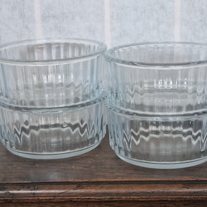 May include: Four clear glass ramekins with vertical ribbed sides are stacked in pairs. The ramekins are round with a slightly flared rim. They sit on a dark wooden surface.