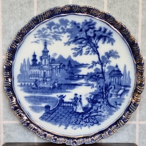 May include: A blue and white porcelain plate with a gold rim. The plate features a detailed scene of a castle and a garden with a bridge and a river.