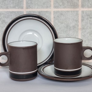 Może przedstawiać: Dwa brązowe ceramiczne kubki do kawy z białym wnętrzem i czarnym wykończeniem. Kubki stoją na pasujących spodkach z białym wnętrzem i czarnym wykończeniem.