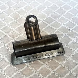 May include: A vintage Waverley clip, made of aged metal, rests on a white, patterned surface. The clip features a cylindrical body, a hinged top with a circular opening, and a rectangular base with the text "WAVERLEY CLIP".