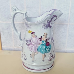 Puede incluir: Una jarra de cerámica blanca con asa y diseño floral. La jarra presenta una escena pintada de dos personas bailando, una con un vestido rosa y la otra con un vestido azul.