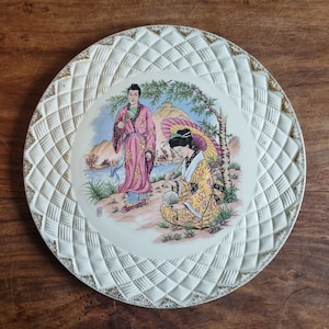 May include: A decorative plate featuring a scene with two figures in traditional Asian dress. The plate has a white, textured border with gold detailing. The central image depicts a landscape with mountains and water.