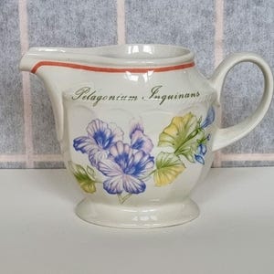 Può includere: Un creamer in ceramica bianca con un design floreale con fiori blu, viola e gialli. Il creamer ha un bordo rosso e il testo "Pelagonium Inguinans" è stampato sul lato.