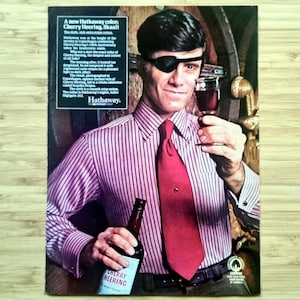 Puede incluir: Anuncio vintage con un hombre con un parche en el ojo, una camisa a rayas rosas y una corbata roja, sosteniendo un vaso de líquido rojo y una botella de Cherry Heering. El anuncio incluye texto de Hathaway.