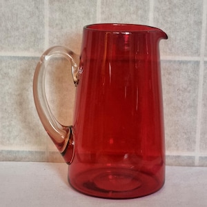Könnte beinhalten: Eine rote Glaskanne mit einem durchsichtigen Glasgriff. Die Kanne ist im traditionellen Stil mit einem breiten Boden und einem schmalen Hals.