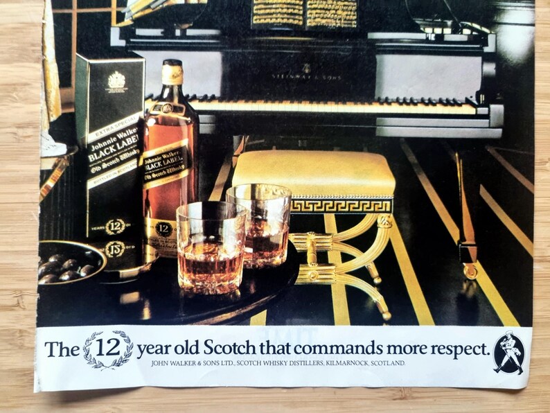 May include: An advertisement featuring a bottle and box of Johnnie Walker Black Label Scotch whisky, two glasses of amber liquid, and a piano. The text reads "The 12 year old Scotch that commands more respect."