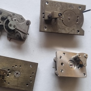 May include: Close-up of several antique watch movement components. The parts are made of brass and silver-colored metal. The components include plates, gears, and levers, showcasing intricate mechanical designs. The image highlights the detailed craftsmanship of the watch parts.