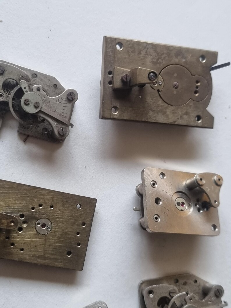 May include: Close-up of several antique watch movement components. The parts are made of brass and silver-colored metal. The components include plates, gears, and levers, showcasing intricate mechanical designs. The image highlights the detailed craftsmanship of the watch parts.
