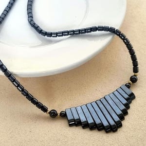 May include: A black beaded necklace with a fan-shaped pendant. The necklace features small, rectangular beads and a gold-colored clasp. The pendant is made of flat, rectangular beads in a dark gray color. The necklace is displayed on a white plate.