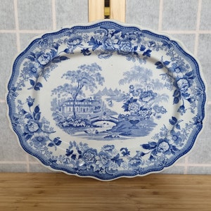 Puede incluir: Un plato ovalado de cerámica azul y blanco con borde festoneado. El plato presenta un diseño floral azul y blanco con una escena central de un puente sobre un río con una casa y árboles en el fondo.