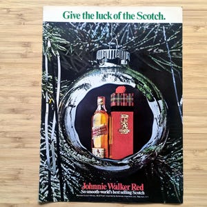 Puede incluir: Un anuncio de Johnnie Walker Red Scotch, que muestra una botella y una caja de regalo dentro de una bola de Navidad. El texto dice "Give the luck of the Scotch." y "So smooth-world's best selling Scotch."