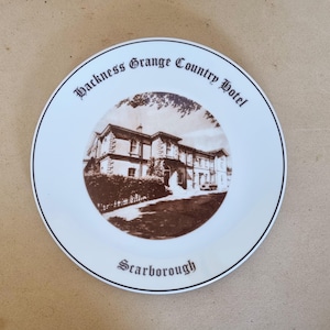 Puede incluir: Plato decorativo blanco con una imagen sepia del Hackness Grange Country Hotel. El plato tiene un borde negro y el texto "Hackness Grange Country Hotel" y "Scarborough".