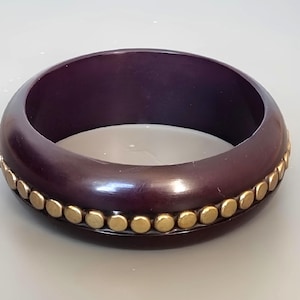 Könnte beinhalten: Ein dunkelvioletter Armreif mit einer Reihe kleiner, goldfarbener Nieten. Das Armband ist dick und hat eine glatte, polierte Oberfläche. Die Nieten sind gleichmäßig am äußeren Rand des Armbands angeordnet.