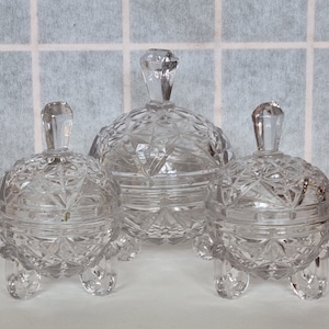 Könnte beinhalten: Drei durchsichtige gläserne Deckelgläser mit einem Diamantenmuster. Die Gläser haben jeweils drei Beine und sind in einer Reihe auf einer weißen Oberfläche angeordnet.