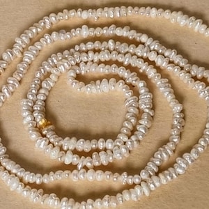 May include: A long strand of white freshwater pearls, strung together to form a necklace. The pearls are irregular in shape and have a slightly iridescent sheen.