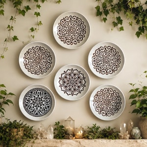May include: A collection of six circular ceramic wall plates arranged in a cluster. Five plates feature intricate brown mandala-style patterns on a speckled cream background, while one plate displays a bold black and white geometric floral design.