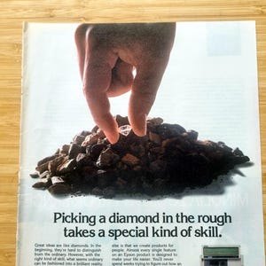 Könnte beinhalten: Eine Anzeige zeigt eine Hand, die einen Diamanten aus einem Haufen roher Steine auswählt. Der Text lautet: „Einen Diamanten im Rohzustand zu finden, erfordert eine besondere Fähigkeit.“