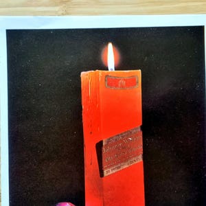 May include: A lit, rectangular red candle with a label that reads "Johnnie Walker Old Scotch Whisky". The candle is set against a black background. The flame is bright and yellow.